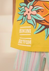 Gul bomuldsbluse med et tropisk design, der har en pink blomst og grønne blade. Teksten "BIKINI BOTTOM" står i hvidt. Stribede stofshorts.