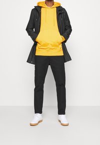 Persona con una sudadera amarilla brillante debajo de una chaqueta negra, pantalones negros y zapatillas blancas, de pie frente a un fondo blanco plano.