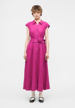 Naine seisab, seljas äravarrukateta fuksia toonis nööpidega kleit, millel on lai vöö ja lillede detail, seljas heledad sokid ja mustad lamedad kingad.