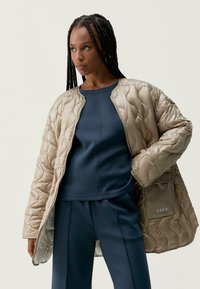 Prošívaná béžová bunda s hladkou texturou, opatřená zipovým zapínáním a boční kapsou, která je navrstvena na tmavě modrém přiléhavém topu a odpovídajících kalhotách.