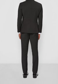 Zwarte op maat gemaakte pak met een enkelbreasted jasje en bijpassende broek. Glad materiaal, minimalistisch ontwerp en glanzende zwarte dresschoenen.