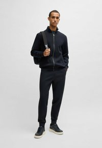 Svart zip-up jacka med hög krage, i kombination med matchande byxor. Har en slät tygkvalitet och ribbade muddar. Bärs med mörka sneakers.