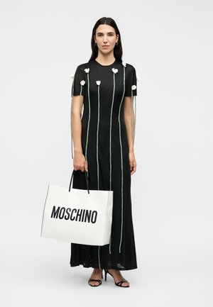 Femme portant une robe noire avec de longues tiges vertes et des fleurs blanches, tenant un grand sac cabas blanc Moschino, debout sur des talons hauts.
