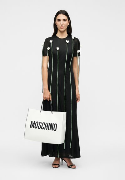 Femme portant une robe noire avec de longues tiges vertes et des fleurs blanches, tenant un grand sac cabas blanc Moschino, debout sur des talons hauts.