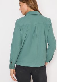 Chemise légère verte à boutons avec col, détail de pli au dos et bouton aux manches. Texture lisse avec une coupe décontractée.