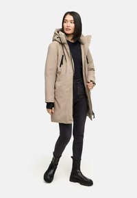 Beige parka med hætte, lynlås lommer og sorte detaljer. Båret med mørke jeans og sorte støvler, med en glat, isoleret tekstur.