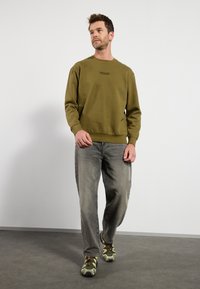 Oliwkowy sweter z okrągłym dekoltem i ściągaczami na rękawach, w połączeniu z jasnoszarymi dżinsami i sneakersami w kamuflażowy wzór w odcieniach zieleni.
