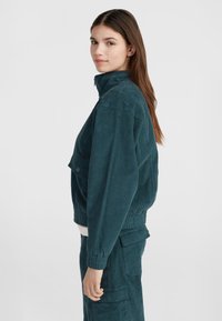 Giacca in corda color teal con colletto alto, vita elastica e tasche laterali. Il modello si posa di lato, evidenziando il tessuto testurizzato.