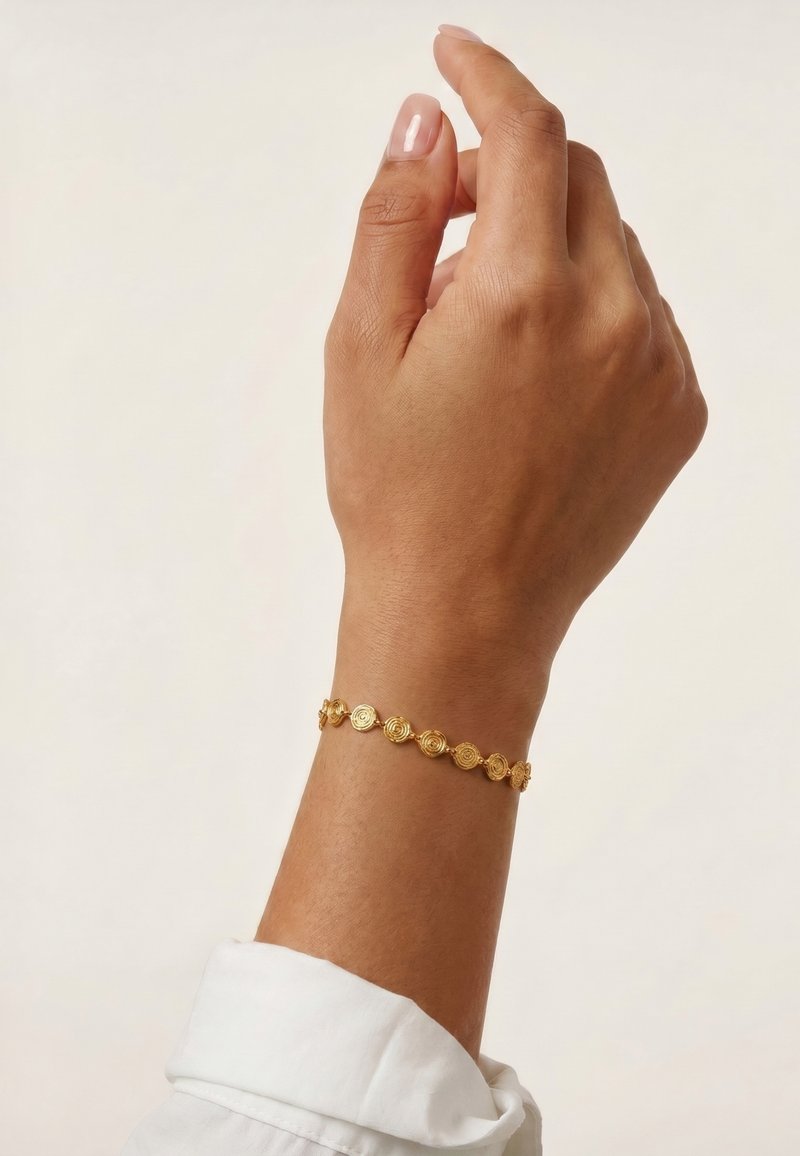 Main à la peau brun moyen portant un bracelet doré à motif circulaire, légèrement relevée avec une manche blanche visible au poignet.