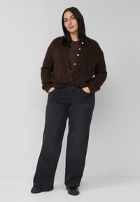 Brązowy sweter na drutach z przodu zapinany na guziki, noszony na ciemnobrązową koszulę, zestawiony z ciemnymi prostymi dżinsami i czarnymi butami.