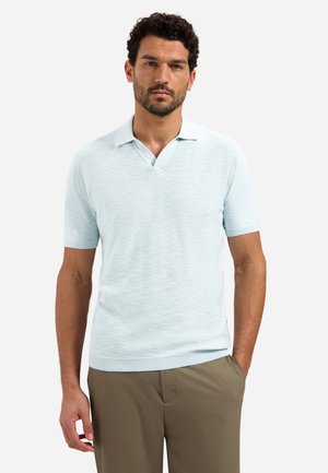 Man met krullend donker haar, gekleed in een lichtblauw, gebreid, polohemd met korte mouwen en een broek in bruine kleur, staand met één hand in zijn zak.