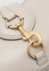 Detalj av en beige läderväska som har en klämma i guldton med en cirkulär ring och två läderremmar. Slät textur och rena stygn.