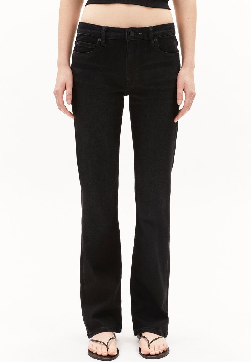 Personne portant un jean noir taille haute à jambes droites, associé à des tongs noires, debout devant un fond blanc uni.