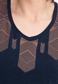 Marinblå bomullst-shirt med geometriskt mönster i ljust rosa på halsringningen och den övre framsidan, med en V-formad krage.