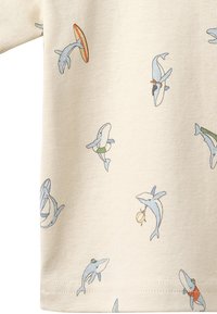 Tissu beige avec des dauphins bleu clair ludiques portant des accessoires comme des chapeaux et tenant des objets tels qu'un ballon de plage et une planche de surf.
