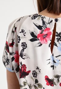 Floral blouse with short sleeves, featuring a cream background, multicolored flowers in red, black, and blue, and a keyhole back detail with button.