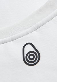 Tela de algodón blanca con una textura suave; presenta un logo negro en forma de aguacate con círculos concéntricos cerca del escote.