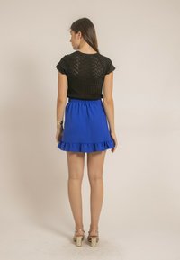 Haut en tricot noir à manches courtes, avec un dos texturé ; associé à une jupe bleue à volants. Le modèle porte des sandales à talons dorées.
