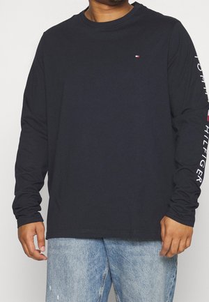 Man draagt een zwart Tommy Hilfiger shirt met lange mouwen, met een logo op de borst en een merktekst op de rechtermouw, gecombineerd met lichtblauwe jeans.