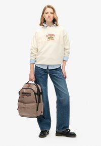 Beige ryggsäck med svarta detaljer, med en framficka, logotyp och röda dragkedjetaggar; bärs med en krämfärgad tröja och blå jeans.