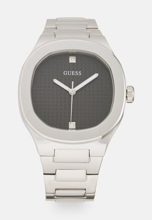 Sølv rustfritt ståluhr med et åttkantet hus, svart teksturert urskive, hvit timeviser og firkantede timemarkører. Merket "GUESS" er vist.