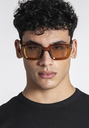 Joven con cabello rizado y oscuro, que lleva gafas de sol rectangulares grandes con lentes de color ámbar y una camiseta negra acanalada, con un pequeño pendiente de aro.