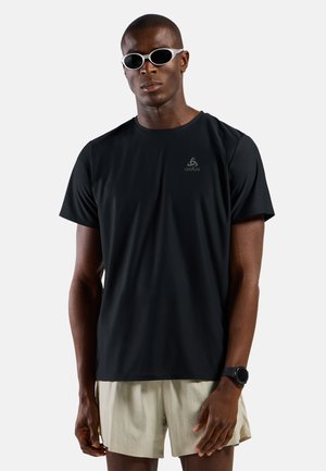 Mann trägt ein schwarzes, kurzärmeliges Sportshirt, beigen Shorts, weiße ovale Sonnenbrille und eine schwarze Smartwatch, steht vor einem schlichten Hintergrund.