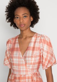 Vestido envolvente em tecido leve com decote em V, mangas curtas bufantes e padrão xadrez rosa e branco. Cinta na cintura.