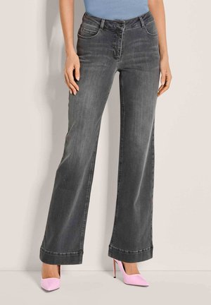 Vrouw draagt hooggetailleerde grijze jeans met wijde pijpen en roze pumps, staand tegen een lichte muur.