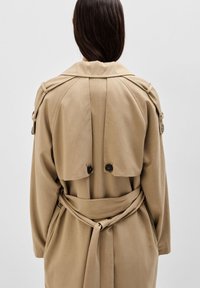 Beżowy płaszcz trench z podwójnym zapięciem, szerokim kołnierzem i wiązanym pasem. Posiada akcenty guzikowe oraz usztywnione plecy z klapą.