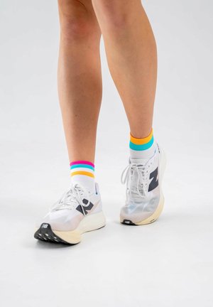 Beine mit weißen Turnschuhen und weißen Socken mit bunten Streifen in Pink, Blau, Gelb und Orange, die auf einem einfachen weißen Hintergrund stehen.