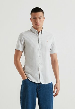 Junger Mann mit kurzen Haaren, der ein kurzärmeliges, hellgrau gestreiftes Hemd mit Knöpfen und blaue Jeans trägt und mit einer Hand in der Tasche steht.