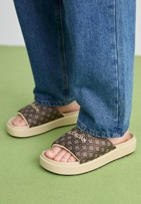 Person iført brune mønstrede slidesandaler med beige såler og blå jeans, som står på et grønt tæppe.
