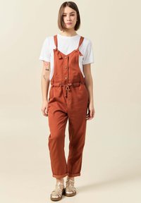 Rostfärgade hängselbyxor med justerbara remmar, knappar framtill och dragsko i midjan; lagrade över en enkel vit t-shirt med korta ärmar.