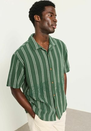 Mann in grünem Kurzarmhemd mit weißen vertikalen Streifen, beigen Hosen, Hände in den Taschen, schaut nach rechts vor einem einfarbigen Hintergrund.