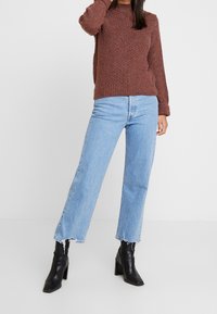 Femme portant un pull en maille marron, un jean court bleu clair à ourlet brut, et des bottines noires à talons, debout devant un fond blanc.
