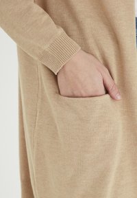 Gilet en tricot beige avec des poignets côtelés et une poche latérale, offrant une texture douce et une coupe décontractée. Une main visible repose dans la poche.