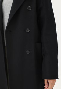 Lång svart ullrock med dubbelknäppning, fyra svarta knappar, slag i kragen och sidoficka. Mjuk textur och skräddarsydd passform.