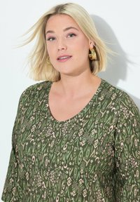Groene blouse met een patroon en een V-halsontwerp, korte mouwen en abstracte vormen in roze en crèmetinten. Glad stofoppervlak.