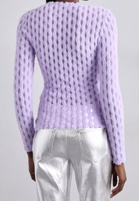 Person som bär en genomskinlig lila texturerad stickad tröja och glänsande silverbyxor, visad bakifrån mot en enfärgad bakgrund.