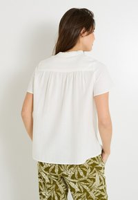 Blouse blanche à manches courtes avec un col à volants et un dos plissé, assortie à un pantalon à motif de feuilles vertes. Tissu texturé.