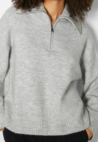 Suéter de punto gris con un alto cuello y cremallera corta en la parte delantera. Presenta puños y dobladillo acanalados, hecho de un material suave y texturizado.