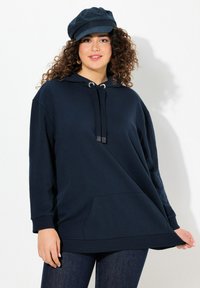 Felpa blu navy con tasca frontale, cappuccio regolabile con coulisse e polsini a costine, abbinata a jeans in denim scuro e un cappellino coordinato.