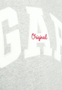 Primer plano de una tela gris con grandes letras blancas y la pequeña palabra bordada en rojo "Original" en un sudadera.