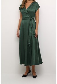 Groene wikkeljurk met korte kapmouwen, een gestrikt detail op de taille, vloeiende stof en een midi-lengte. Gekleed met zwarte puntige hakken.