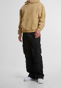 Muž oblečený v béžové mikině s kapucí, černých cargo kalhotách s velkými kapsami a bílých teniskách stojící proti jednobarevnému světlému pozadí.