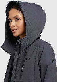 Veste grise courte avec une grande capuche, dotée de cordons de serrage, d'une texture lisse et d'un logo sur la manche. Le design met l'accent sur la fonctionnalité.