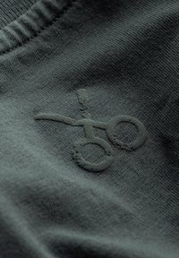 Tessuto verde scuro con un logo a forbice ricamato e testurizzato. Texture liscia con un design leggermente in rilievo.