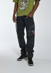 Jeans de mezclilla negra con un corte holgado, que presentan un gráfico detallado de un gato negro y acentos rojos en el muslo. Combinados con zapatillas altas negras.