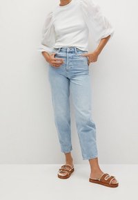 Helle blaue hochgeschnittene Jeans mit geradem Schnitt, kombiniert mit einem weißen Oberteil mit texturierten Puffärmeln und braunen Sandalen mit zwei Riemen.